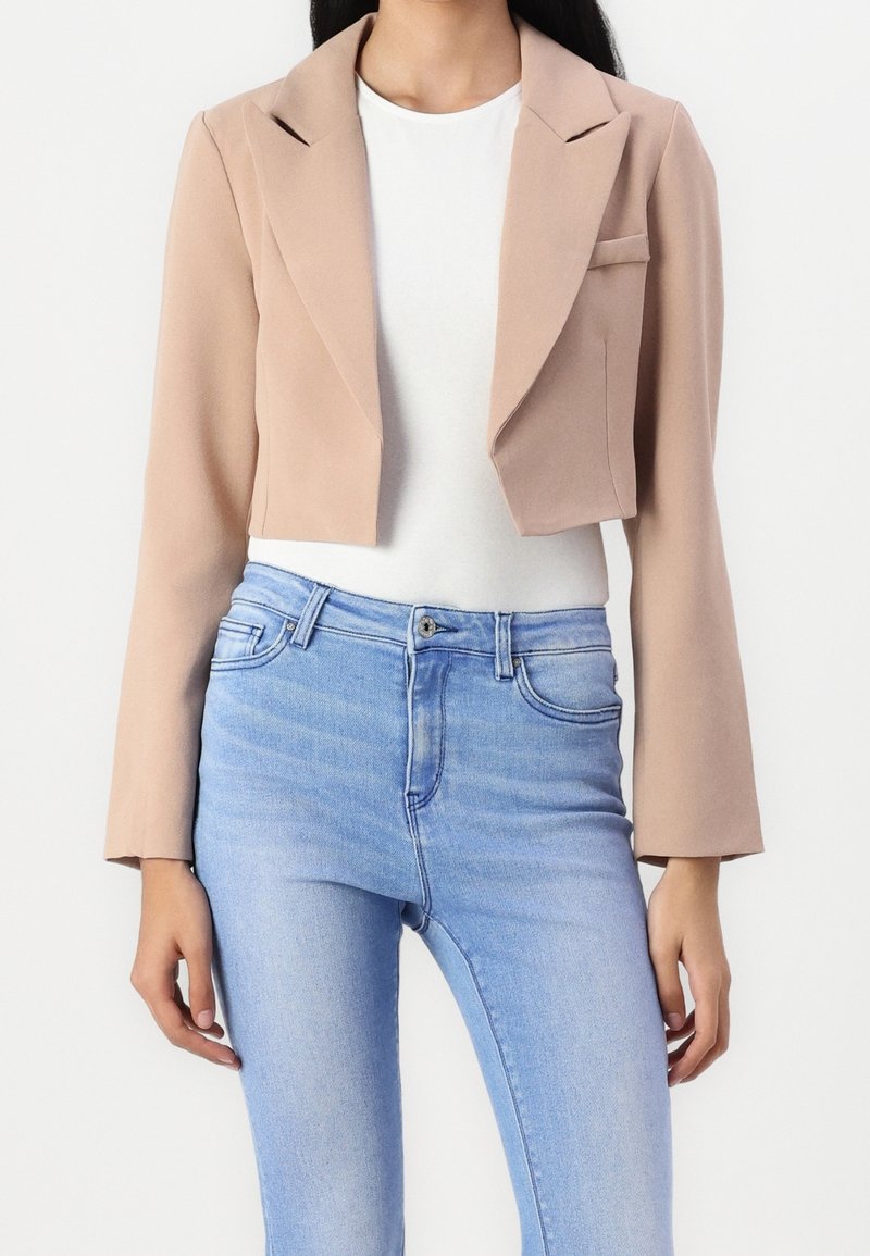 Femme portant un blazer beige court sur un top blanc avec un jean skinny bleu clair, debout devant un fond blanc uni.
