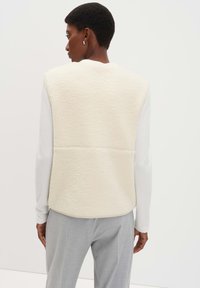 Cremfarbener Fleece-Weste mit strukturierter Oberfläche, kombiniert mit einem weißen Langarmtop und hellgrauen Hosen. Einfaches, modernes Design.