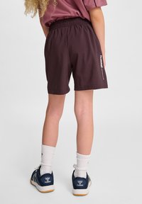 Mörkröda shorts med elastisk midja, sidficka och vit logodetalj på vänster sida. Bärs med vita strumpor och mörkblå sneakers.