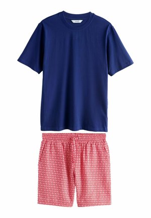 Mann mit dem Rücken zur Kamera, trägt ein schlichtes dunkelblaues kurzärmeliges Shirt und rote gemusterte Shorts, die Hände in den Taschen, vor einem einfarbigen Hintergrund.