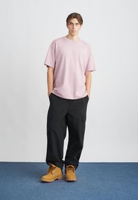 Νέος άνθρωπος στέκεται με τα χέρια στις τσέπες, φορώντας θεαματική μοβ oversize μπλούζα, μαύρο cargo παντελόνι και καφέ μποτάκια με κορδόνια σε μπλε δάπεδο.