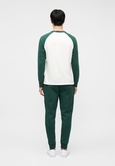 Άνδρας που στέκεται με την πλάτη προς τα εμπρός, φοράει λευκό και πράσινο μακρυμάνικο πουκάμισο, σκούρο πράσινο παντελόνι και λευκά αθλητικά παπούτσια σε μονόχρωμο φόντο.