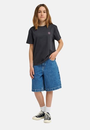Dziewczyna stoi z ręką w kieszeni, ubrana w czarny t-shirt, niebieskie dżinsowe spodenki sięgające do kolan, białe skarpetki i czarne trampki.