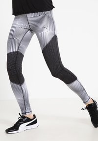 Sort-hvide leggings med mønster, der har geometriske designs, strukturerede sorte detaljer og en tætsiddende silhuet, parret med sorte træningssko.