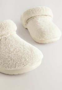 Zapatillas de felpa blanca con punta redondeada, textura suave y puño doblado. Suela de goma duradera y de color claro.