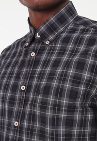 Camicia a maniche corte a quadri in blu navy, grigio e bianco. Presenta un colletto abbottonato e una tasca frontale. Tessuto di cotone morbido con bottoni a contrasto.
