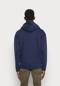 Man som bär en marinblå huvtröja och olivgröna byxor, stående vänd mot en enkel ljusgrå bakgrund.