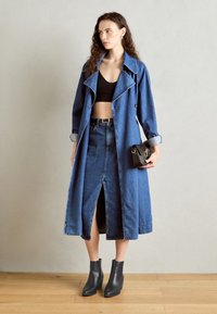 Cappotto trench in denim con una vestibilità leggermente oversize, caratterizzato da un design doppiopetto, ampio colletto e uno spacco frontale. Abbinato a stivali neri alla caviglia.