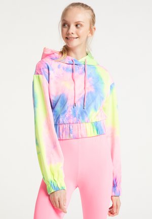Młoda kobieta ubrana w kolorową krótką bluzę z kapturem w stylu tie-dye i jasnoróżowe legginsy, patrzy na bok z lekkim uśmiechem.