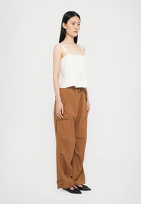 Weißes Cropped-Top mit dünnen Trägern und einem Rüschensaum, kombiniert mit braunen Cargo-Hosen mit Seitentaschen und verstellbaren Bündchen. Schwarze spitze Ballerinas.