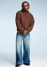 Ung man med flätat hår iförd en brun hoodie och vida blekta blå jeans, stående med händerna hopknäppta framför sig.