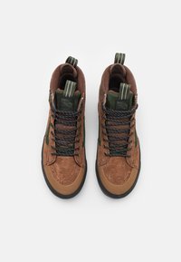 Bruna och gröna högskor med svarta och vita snören, dragflikar och svarta gummisulor, visade uppifrån på en vit bakgrund.