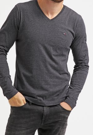Homme portant un t-shirt à manches longues gris foncé avec col en V et petit logo sur la poitrine, et un jean noir, mains partiellement dans les poches.