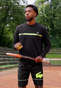 Sweatshirt preto com riscas verdes e azuis, calções pretos com um logotipo de crocodilo neon verde, segurando uma bola de ténis e uma raquete.