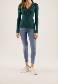 Haut à manches longues en maille côtelée turquoise avec un col en V et un détail avant tordu, associé à un jean bleu slim et des baskets blanches.