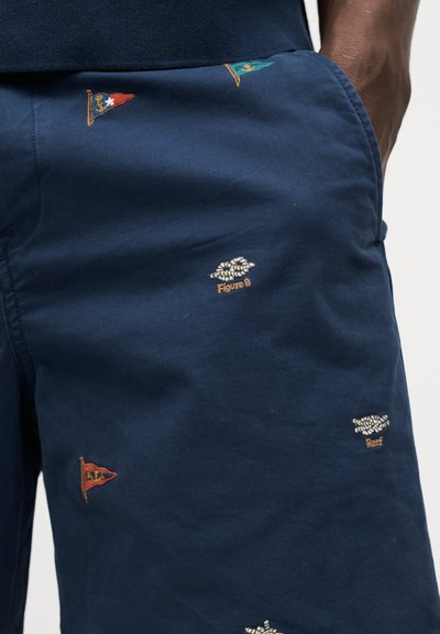 Σορτς σε ναυτικό μπλε με μικρές κεντημένες σημαίες και ναυτικά σχέδια, φορεμένα από άτομο με σκουρόχρωμο δέρμα και χέρι στην τσέπη.