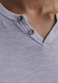 Camisa henley de color púrpura claro con una superficie texturizada, con un cuello en V y dos botones en el cuello. Tejido suave y diseño casual.