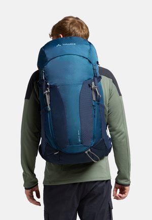 Zaino blu e navy scuro con spallacci regolabili, tasche laterali in rete e patta superiore. Caratterizzato da accenti riflettenti e da una forma aerodinamica.