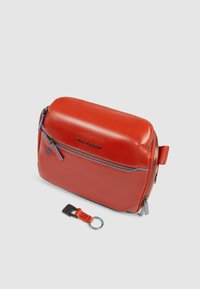 Bolso bandolera de cuero rojo con textura suave, acentos de cremallera azul y un llavero desmontable. Cuenta con una forma rectangular y un asa lateral.