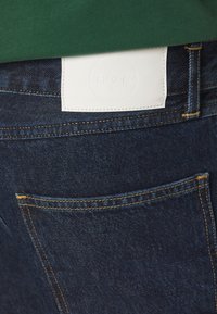 Mörka denimjeans med en texturerad finish, med guldtråd, en bakficka och en vit läderlapp med präglad text.