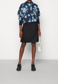 Stickad marinblå tröja med färgglada blommönster, matchad med en svart kjol. Modellen håller en krämfärgad handväska och bär svarta platåskor.