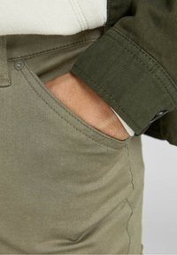 Hand delvis i framfickan på ljus olivgröna byxor, bärande en mörk olivgrön jacka med synlig manschett och knapp.