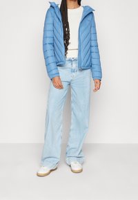 Světlá modrá péřová bunda s vertikálním prošíváním, kapucí a zipem, v kombinaci s světle modrými džínami s vysokým pasem a bílými teniskami.