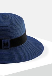 Niebieski pleciony kapelusz z szerokim rondem i czarną tkaniną z wypukłą literą "M" jako detal. Tekstura powierzchni i zdefiniowany fason.