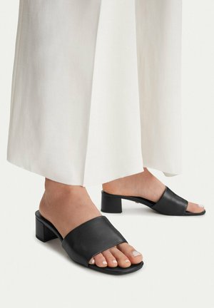 Pies con sandalias negras de tacón bloque y punta abierta, combinadas con pantalones blancos largos de pierna ancha sobre un fondo liso.