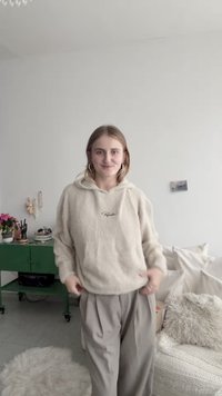 Smėlio spalvos fliuso džemperis su kapišonu, priekyje kišenė ir siuvinėtas tekstas, derinamas su šviesiai pilkomis plačiomis kelnėmis. Minkšta tekstūra ir laisvas kirpimas.