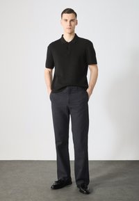 Zwarte gebreide poloshirt met korte mouwen en drie knopen, gedragen met donkere broek en zwarte schoenen op een grijze vloer.