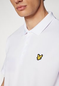 White polo shirt with a classic collar, featuring a black and yellow embroidered bird logo on the chest. Smooth, lightweight fabric.