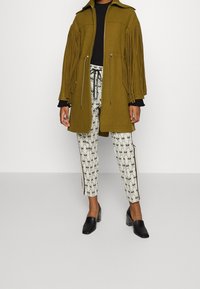 Olivgrön fransad kappa med fickor med dragkedja, matchad med mönstrade vita byxor med svarta mönster och sidan ränder, samt svarta skor med blockklack.