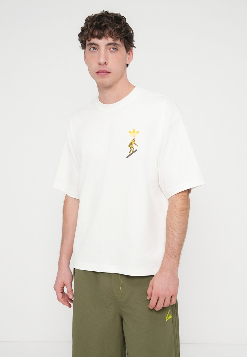 Vitt bomulls-T-shirt med korta ärmar, med en gul grafik av en skidåkare och logotyp på vänstra bröstet. Kombinerad med olivgröna shorts.