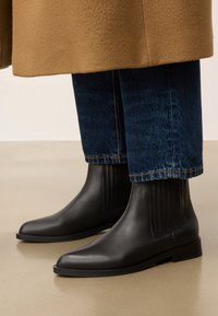 Czarne skórzane botki za kostkę z elastycznymi bocznymi panelami, kwadratowym noskiem i niskim czarnym obcasem. Noszone z niebieskimi dżinsami i jasnobrązowym płaszczem.