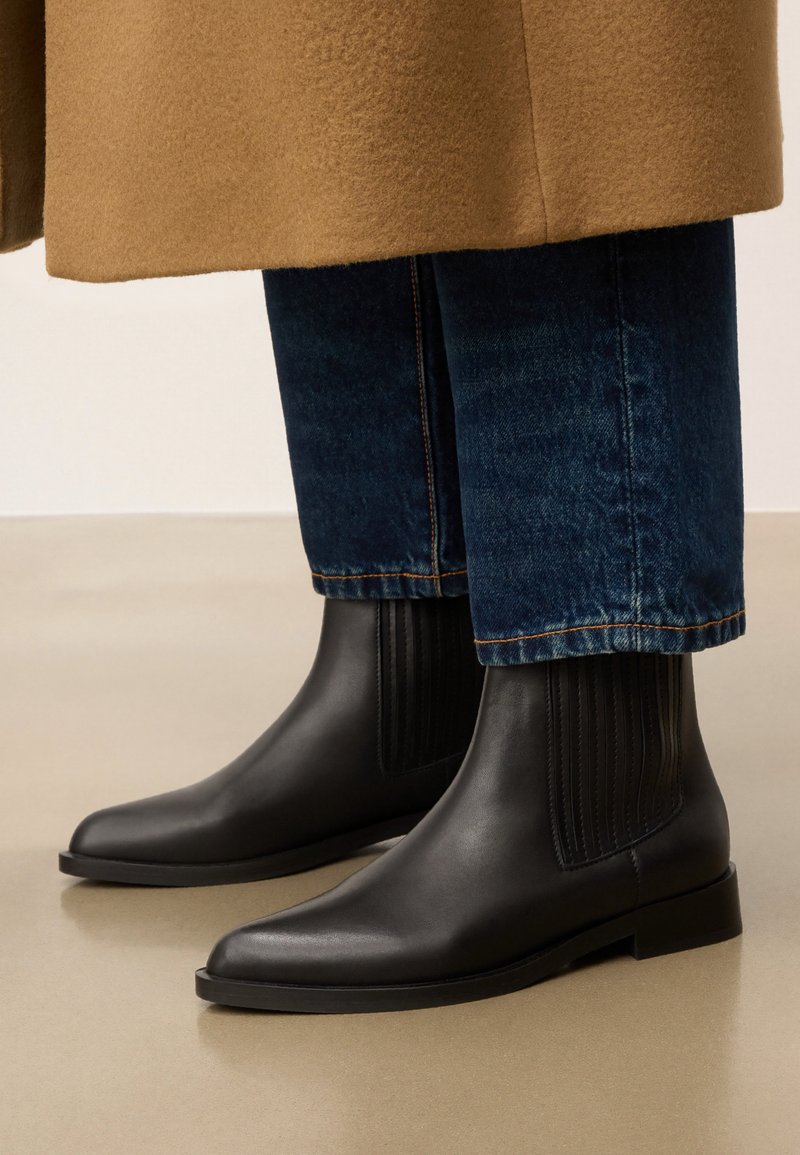 Czarne skórzane botki za kostkę z elastycznymi bocznymi panelami, kwadratowym noskiem i niskim czarnym obcasem. Noszone z niebieskimi dżinsami i jasnobrązowym płaszczem.