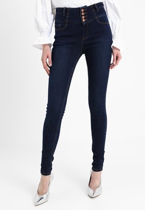 Vrouw draagt donkerblauwe high-waisted skinny jeans met vier knopen en witte blouse met lange mouwen, gecombineerd met zilveren puntige hoge hakken.