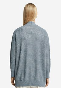 Ljusblå tröja med en texturerad hexagonal mönstring, ribbade muddarna och en något oversized passform. Bakvyn framhäver den intrikata designen.