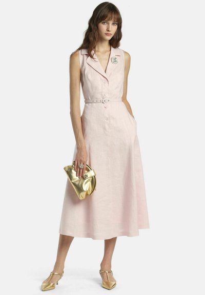 Žena v bezrukávových bledoružových midi šatách so zaväzovaním v páse, drží zlaté kabelku a nosí zlaté špicaté lodičky a prsteň.