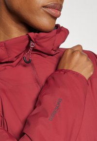 Veste isolée bordeaux avec un col haut, fermeture éclair, cordon de serrage réglable et un logo "Didriksons" sur la manche. Texture lisse.