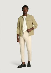 Beige Zip-Jacke über einem hellen Hemd, kombiniert mit cremefarbenen Hosen und schwarzen Schuhen. Die Jacke verfügt über eine Brusttasche und hat eine glatte Textur.