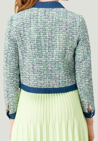 Teksturowana, tkana kurtka w kolorach zielonym, białym i niebieskim z kołnierzem z denimu. Jasna plisowana, miętowo-zielona spódnica dodaje kontrastu. Rękawy 3/4 z mankietami.
