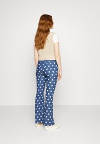 Colete bege tricotado sobre uma t-shirt branca, combinado com calças azul flare com padrões florais brancos. Calçado casual completa o visual.