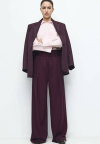 Bordeaux kleurig colbert over een lichtroze overhemd, gecombineerd met hoge taille en wijd uitlopende broekspijpen. Soepele textuur, op maat gemaakt ontwerp en knoopdetails.