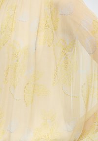 Διάφανο μπεζ ύφασμα με κίτρινα και ανοιχτομπλε λουλουδάτα σχέδια και απαλά πτυχώματα.