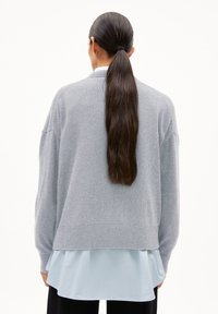 Grauer, übergroßer Pullover mit geripptem Saum und abgesenkten Schultern, getragen über einem hellblauen, mehrlagigen Oberteil. Der Pullover hat einen lässigen Schnitt und eine weiche Textur.