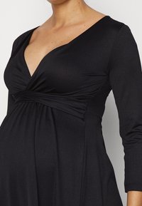 Svart klänning med djup V-ringning, långa ärmar och rynkad midjedetalj. Tillverkad av en mjuk, elastisk tyg. Elegant drapering och figurnära silhuett.