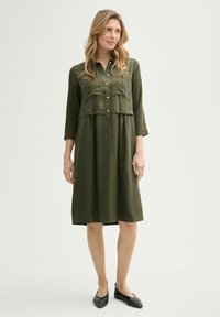 Olivgrünes Kleid mit durchgehender Knopfleiste, zwei Brusttaschen und Dreiviertelärmeln. Weicher Stoff mit lockerer Passform, knielanges Design.