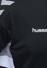 Svart atletisk tröja med en slät yta, grå detaljer och vit "hummel"-logotyp placerad på framsidan. Reflekterande detalj på axeln.