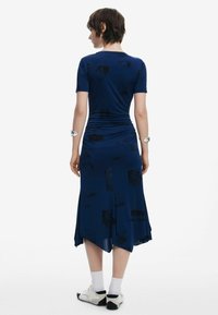 Marinblå klänning med en figurnära bodice, korta ärmar, rynkad midja och en svängd kjol med ett subtilt svart mönster. Nederkanten är asymmetrisk.